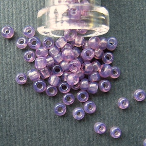 May include: A collection of translucent, lavender-colored seed beads spilling from a clear glass container. The beads are small, round, and have a hole running through the center, ideal for jewelry making or crafts.