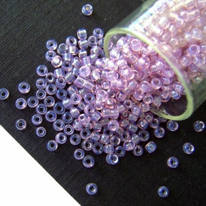 May include: A collection of translucent, lavender-colored seed beads spilling from a clear glass container onto a black surface. The beads are small, cylindrical, and have a hole running through the center, ideal for jewelry making and crafts.