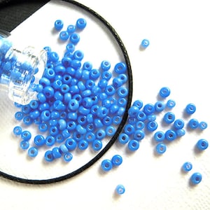 May include: A small glass jar filled with light blue seed beads. The beads are spilling out of the jar onto a white surface. There is a black cord wrapped around the jar.
