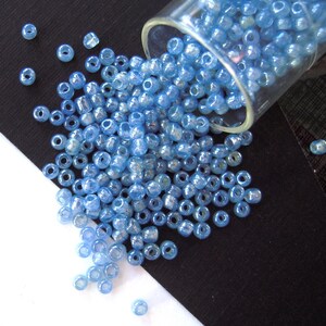 May include: A glass jar filled with light blue iridescent seed beads, spilling out onto a black surface. The beads are small and round, with a shiny finish.
