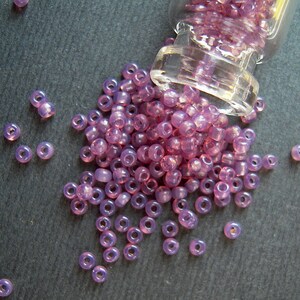 May include: A glass jar filled with small, iridescent purple beads. The jar is tipped over, spilling the beads onto a dark gray surface.