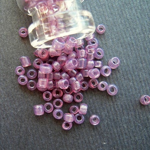 May include: A glass jar filled with small, translucent purple beads. The beads are scattered on a gray surface.