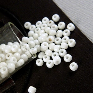 May include: A glass jar filled with white seed beads, spilling out onto a black surface. The beads are small and round, with a smooth, glossy finish.