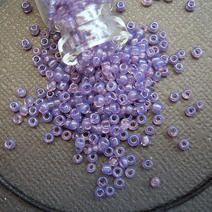 May include: A glass jar filled with small, light purple glass beads. The beads are scattered on a dark gray surface.