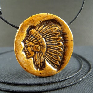 May include: A round, tan-colored pendant featuring a detailed relief of a Native American chief in profile, wearing a feathered headdress. The pendant is strung on a black cord, creating a necklace. The pendant has a speckled texture.