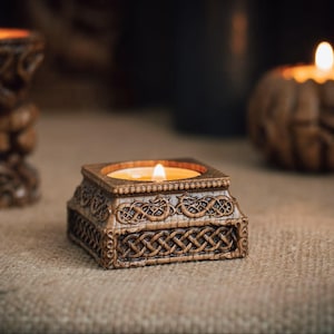 Pode incluir: Um suporte de vela quadrado castanho com padrões de nós esculpidos intrincados nos lados, apresentando uma vela de chá acesa no centro. O suporte tem um acabamento texturizado e está posicionado em primeiro plano com outras duas velas acesas.