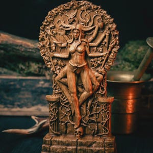 Può includere: Un intaglio in rilievo in legno marrone raffigurante una figura dalle braccia multiple con capelli fluenti, in piedi sopra un teschio e una base in pietra. La figura è circondata da fogliame e viticci intricati, tenendo vari oggetti nelle mani.