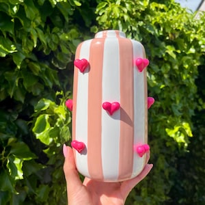 May include: A cylindrical ceramic vase featuring vertical stripes in alternating shades of soft pink and white. The exterior surface is decorated with several small, raised, three-dimensional bright hot pink heart-shaped embellishments.