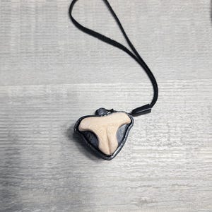 May include: A pendant necklace featuring a light beige, tooth-shaped charm with a dark gray border. The charm is suspended from a black cord. The background is a light gray wooden surface.
