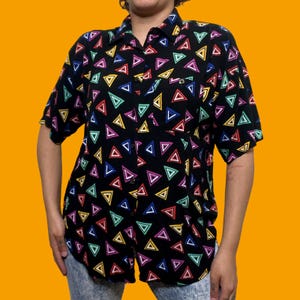 May include: Black button-down shirt with a colorful triangle pattern. The short-sleeved shirt features a pocket on the chest and a collar. The triangles are in various colors, including red, yellow, blue, green, and purple.
