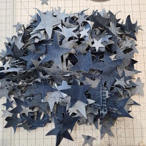 May include: A pile of denim star-shaped fabric cutouts in various shades of blue and gray. The stars are scattered on a cutting mat with grid lines and the Alto's logo. The stars vary in size and are made from recycled denim.