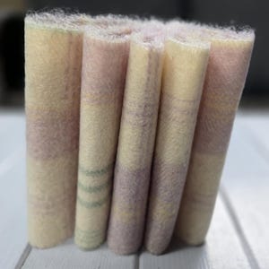 May include: Close-up of a stack of folded fabric. The fabric has a soft, textured appearance with a colour palette of pale yellow, lavender, and light green stripes. The edges of the fabric are slightly frayed.