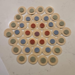 May include: A collection of beige felt circles, each with a smaller, contrasting felt circle in the center. The center circles are in various colors, including green, blue, and red. The arrangement suggests a craft or game component.