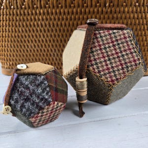 May include: Two handmade, hexagonal pincushions with a patchwork design. One has a button closure and the other has a leather strap with wooden beads. The fabrics include tweed and plaid patterns in shades of brown, green, and red.