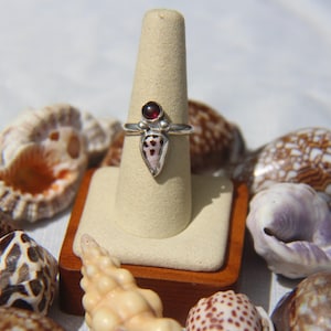 Puede incluir: Un anillo de plata con una gema roja y un diseño único con patrones. El anillo se exhibe en un soporte, rodeado de varias conchas marinas. El diseño del anillo es el punto focal, mostrando sus intrincados detalles y artesanía.