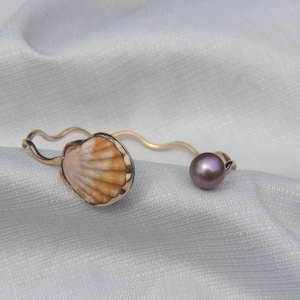 Puede incluir: Una pulsera abierta de tono dorado con un diseño de banda ondulada. Un extremo de la pulsera está adornado con una pequeña concha natural en forma de abanico de color naranja y blanco, mientras que el otro extremo termina en una perla redonda, lisa y de color morado oscuro.
