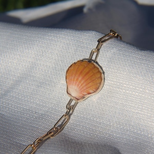 Puede incluir: Una pulsera de cadena en tono dorado que presenta un dije central de concha de abanico. La concha muestra tonos naturales de naranja, bronceado y blanco con crestas radiales. La cadena está compuesta por eslabones metálicos rectangulares.