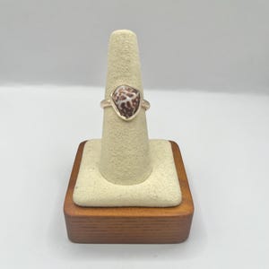 May include: A rose gold ring featuring a natural seashell centerpiece. The seashell has a unique pattern of brown and white spots. The ring is displayed on a beige ring holder with a wooden base, set against a white background.