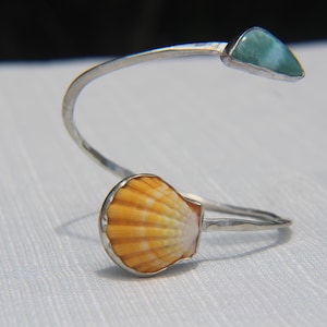 Puede incluir: Una pulsera de puño plateada con una concha natural y una gema triangular azul claro. La concha es naranja y blanca. La pulsera tiene un diseño curvo.