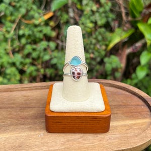 May include: Two silver rings with distinctive stone settings are presented on a ring holder. One ring features a light blue stone, the other a brown and white patterned stone. The rings are displayed on a wooden stand.