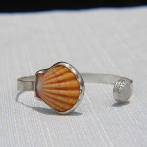 May include: A silver cuff bracelet featuring a seashell design. The seashell is orange and white with a silver border. The bracelet has an open design with a small, round, textured silver end piece.