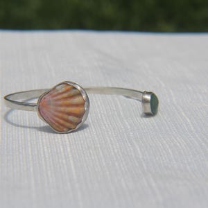 Op de afbeelding: Een zilveren armband met een geschulpte schelp met oranje en witte ribbels aan het ene uiteinde en een kleine, gladde, ronde groene steen in een zilveren zetting aan het andere uiteinde, wat zorgt voor een open ontwerp met een gepolijste afwerking.