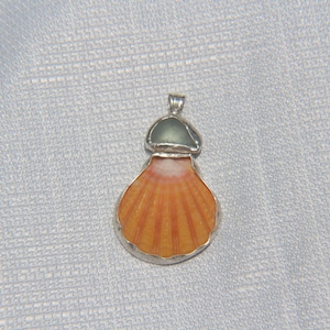 May include: A pendant featuring a fan-shaped orange scallop shell with a white center, topped with a small, smooth, translucent sea-glass piece in a muted sage green. The two elements are encased in a silver-toned metal frame with a bail.