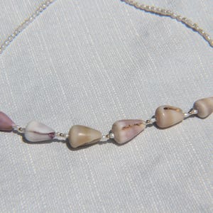 May include: A necklace featuring a strand of small white seed beads interspersed with six smooth, teardrop-shaped seashells in shades of light tan, beige, and soft mauve. The shells are arranged along the lower portion of the delicate beaded chain.