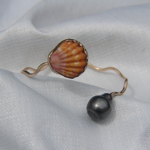 Può includere: Un bracciale aperto color oro con un design a fascia ondulata. Un'estremità del bracciale è decorata con una piccola conchiglia arancione striata, mentre l'estremità opposta termina con una singola perla sferica liscia di colore grigio scuro.