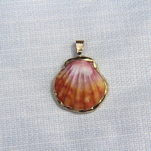 May include: A seashell pendant with a gold-colored frame and bail. The shell is painted with shades of orange, red, and white. The pendant is likely made of ceramic or porcelain and is a piece of jewelry.