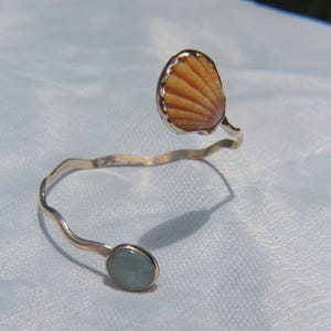 May include: A silver-coloured cuff bracelet with a natural seashell and a small oval-shaped, pale green stone. The seashell is a warm, light orange colour. The bracelet has a wavy design.