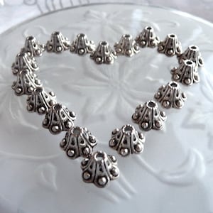 May include: A collection of silver metal bead caps arranged in a heart shape on a white surface. The bead caps have a decorative design with raised dots and a conical shape.