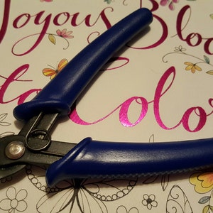 May include: Blue handled wire cutters with black metal jaws. The cutters are on a white background with a pink floral design and the text "Joyous Blooms to Color".