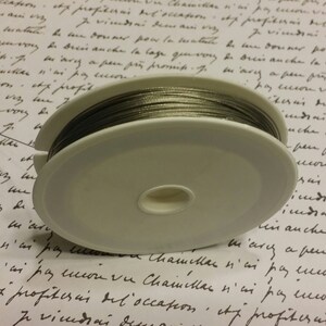 May include: A spool of silver-colored wire on a white plastic spool. The spool is sitting on a piece of paper with handwritten text.