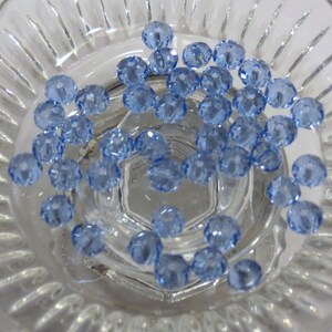 May include: A close-up of a bowl filled with light blue glass beads. The beads are faceted and have a shiny finish.