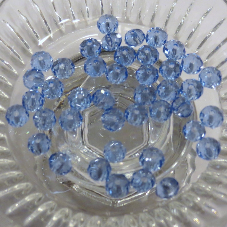 May include: A close-up of a bowl filled with light blue glass beads. The beads are faceted and have a shiny finish.