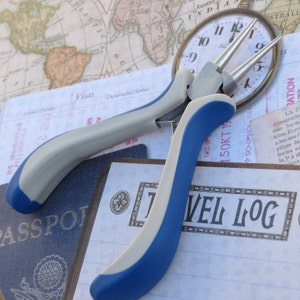 May include: Blue and silver jewelry pliers with a blue handle and a silver handle. The pliers are lying on a white surface with a travel log book and a vintage map in the background.
