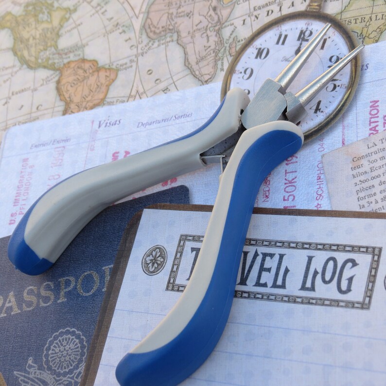 May include: Blue and silver jewelry pliers with a blue handle and a silver handle. The pliers are lying on a white surface with a travel log book and a vintage map in the background.