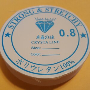 May include: A white spool of 0.8 mm strong and stretchy elastic thread labeled "Strong & Stretchy" with a blue diamond logo and the text "Crysta Line" in Japanese and English. The spool also says "Polyurethane 100%".
