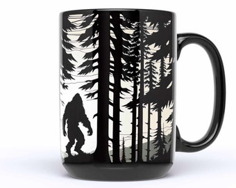 Può includere: Tazza da caffè in ceramica nera con una silhouette bianca di Bigfoot che cammina attraverso una foresta. La tazza ha un grande manico.