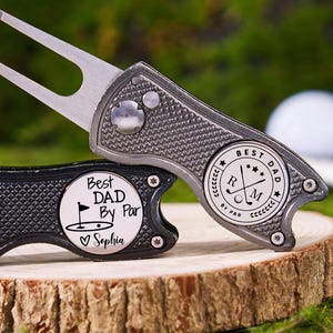 Peut inclure: Deux outils de réparation de divots de golf argentés et noirs, posés sur une surface en bois. L'un porte l'inscription "Best Dad By Par" et un nom gravé. L'autre affiche "Best Dad" et des symboles de golf. Des balles de golf sont en arrière-plan.