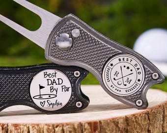 May include: Two golf divot tools with a silver and black finish, resting on a wooden surface. One tool has "Best Dad By Par" and a name engraved. The other tool has "Best Dad" and golf-related symbols. Golf balls are in the background.
