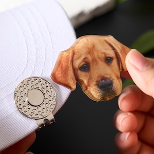 May include: A white golf hat with a silver magnetic hat clip and a golden retriever dog ball marker. The dog marker is a detailed, realistic image of a Labrador Retriever's face.