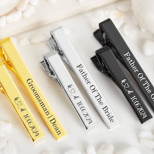 Può includere: Quattro fermagli per cravatta con diverse incisioni. I fermagli per cravatta sono dorati, argentati e neri. Le incisioni includono "Father of the Groom", "Father of the Bride", "Groomsman | Dean" e "kd 10.06.2024".