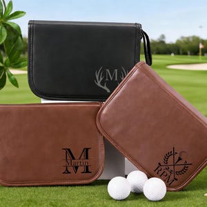 Op de afbeelding: Drie gepersonaliseerde golfaccessoire-etuis in zwart en bruin leer worden gepresenteerd op een groene ondergrond met golfballen en tees. De etuis hebben aangepaste initialen en ontwerpen, waaronder een hertenkop, initialen en een golf-thema-embleem.