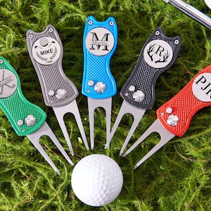 May include: Five personalized golf divot tools in green, gray, blue, black, and red, each with a unique design and initials. A white golf ball and a tee are also visible, all set against a green, mossy background.