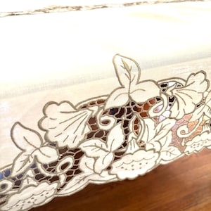 May include: Ivory-colored tablecloth with a detailed floral lace border. The lace features a delicate leaf and flower design, creating an elegant and classic look. The tablecloth is set on a wooden surface, adding warmth to the overall aesthetic.