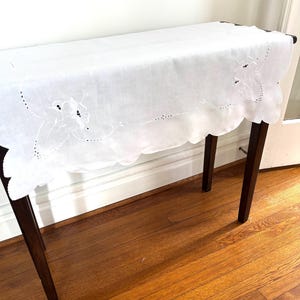 May include: A white rectangular table runner featuring delicate white floral embroidery and cutwork details along the edges. The runner has a scalloped border and is draped over a dark wooden console table, showcasing intricate needlework patterns.