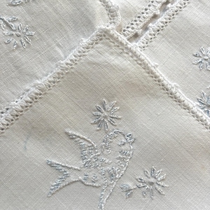 May include: A white fabric textile featuring delicate white embroidery of a bird in flight surrounded by small floral motifs. The edges of the fabric are finished with a decorative white crocheted border, creating a textured and detailed appearance.