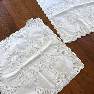 May include: Two white, square, vintage-style pillowcases with a patchwork design. Each pillowcase features a combination of solid white squares and squares with intricate crochet detailing. The edges are finished with a scalloped crochet trim.